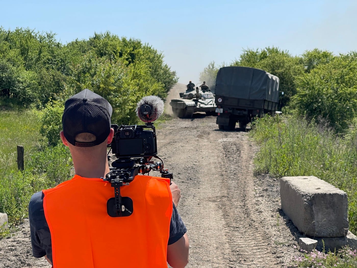 Kameraman natáčí tank a vojenský náklaďák blížící se po prašné cestě.